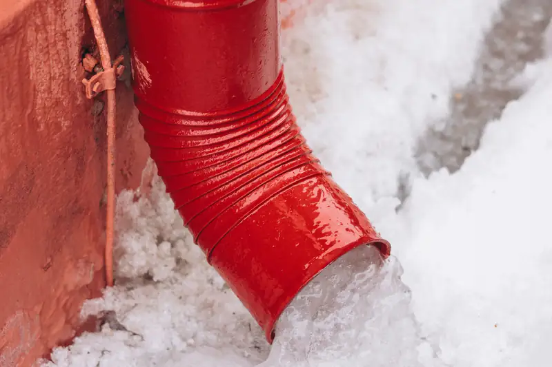 Red downspout surrounded by melting snow next to a house wall