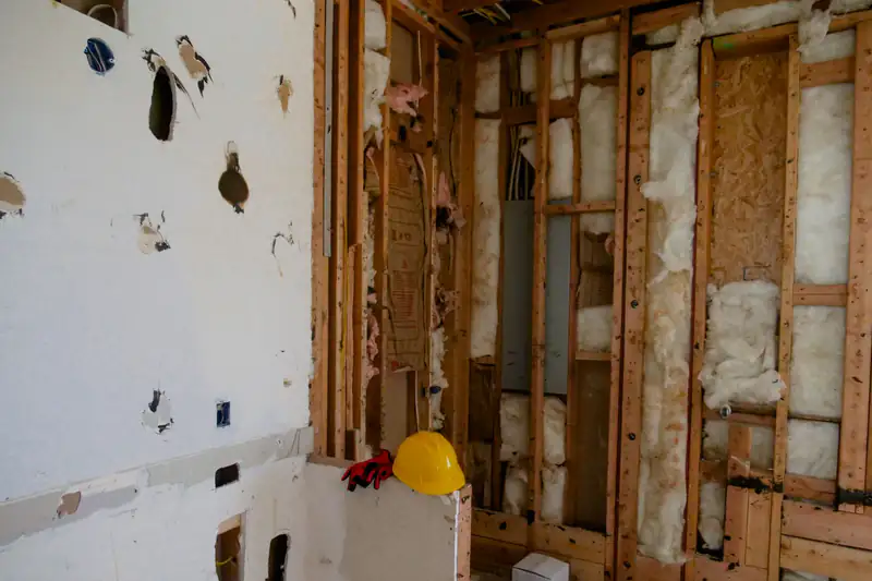 Room with drywall partly removed, exposing wood studs and insulation, and a hard hat on the floor