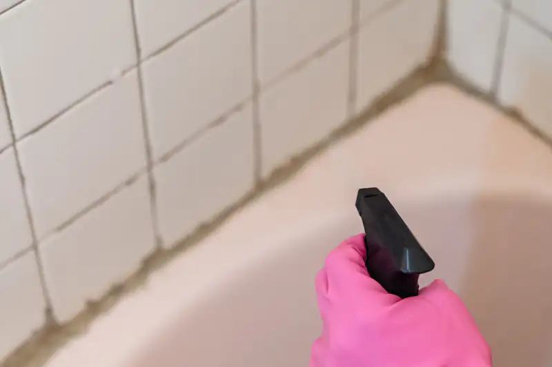 Hand in a pink rubber glove spraying cleaner on bathroom wall tiles